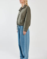 Olive green Murphy bomber jacket with elasticated hem, zip front, and silver snap details worn with blue jeans.
