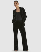 Model wearing black stretch velvet pants with a slight flare leg profile and matching velvet blazer.