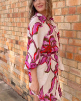 Novara linen-blend shirt dress with a pink and red floral print, button front, and waist tie against a brick wall.