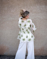 Woman wearing a sage green palm print Tencel-viscose blouse with ruffled details and white wide-leg pants.