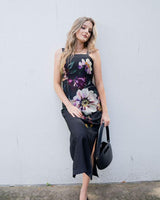Model wearing a black sleeveless Providence midi dress with a bold floral print, square neckline, and side leg slit.