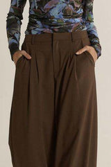 Chocolate brown relaxed-fit trousers with front pleats, side pockets, and a tailored zip fly closure.