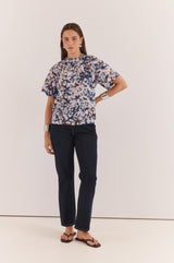 Anika Frill Neck Blouse in a blue abstract print with a gathered neckline and short sleeves, styled with dark denim jeans.