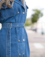 Sleeveless blue denim dress featuring a double-breasted button front, a matching tie waist belt, and side pockets.