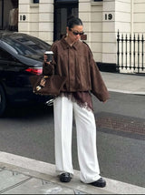 Oversized brown suede-like bomber jacket with a zip front, collar, and elasticated hem worn with white wide-leg trousers.