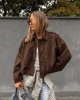 Woman wearing a brown suede bomber jacket with a zip-front, stand-up collar, and relaxed fit against a grey wall.