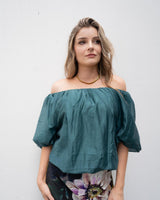 Woman wearing a teal off-shoulder top with a bubble hem and sleeves, styled with a floral skirt and gold necklace.