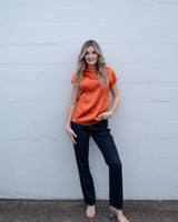 Woman wearing a burnt orange Zafina pleated draped top with cap sleeves and a subtle sheen, paired with dark blue jeans.