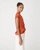 Burnt orange draped top with cap sleeves and a subtle sheen, styled with white tailored pants.