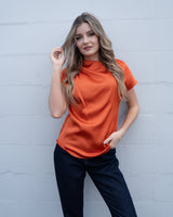 Burnt orange pleated draped top with cap sleeves and a subtle sheen, worn with dark denim jeans.