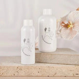Two white bottles of Evella.Co Foaming Bath Bubbles with minimalistic black line art design, placed on beige stone surfaces with a soft floral background.