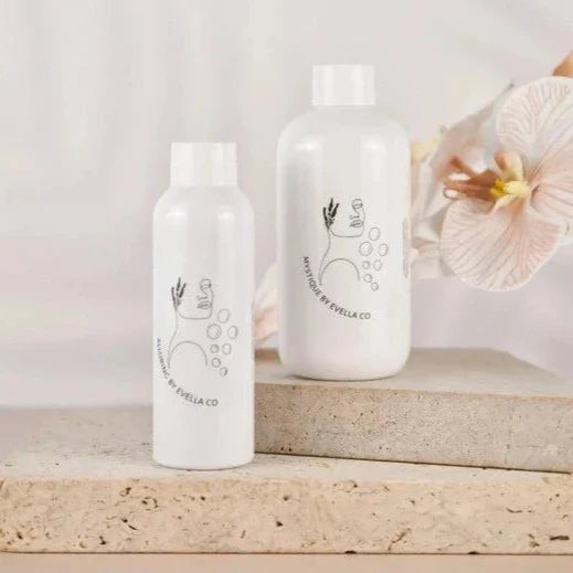 Two white bottles of Evella.Co Foaming Bath Bubbles with minimalistic black line art design, placed on beige stone surfaces with a soft floral background.